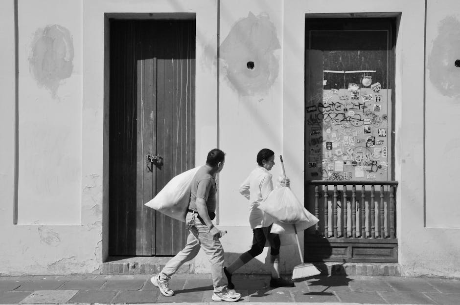 Two street cleaning workers carry bags past a graffiti-covered wall in black and white.